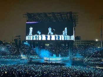 Escenario de concierto nocturno con una gran pantalla LED y miles de espectadores con luces azules y verdes en las gradas
