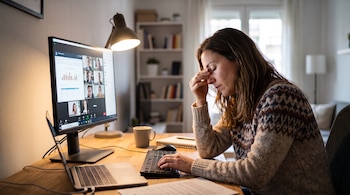 Una mujer sentada a un escritorio se toca los ojos frente a un monitor que muestra una videollamada.