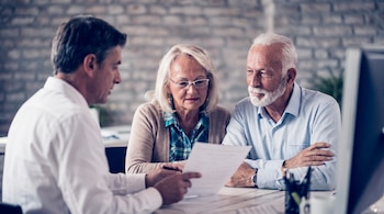 Un asesor financiero explica documentos a una pareja de adultos mayores en una oficina con pared de ladrillo. El hombre mayor tiene barba blanca.