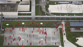 Vista aérea de un patio de prisión. Reclusos en uniformes naranjas y blancos en canchas de baloncesto y una zona de arena, con edificios de prisión y vallas