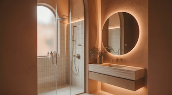 Un baño moderno en tonos tierra con lavabo de travertino, espejo circular retroiluminado y ducha de cristal bajo ventana arqueada, junto a un mueble de madera.