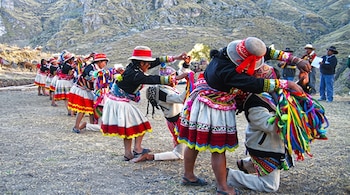 Hombres arrodillados y mujeres de pie con trajes coloridos, sombreros y cintas multicolores en un terreno de tierra junto a montañas.