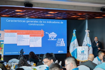 Un hombre con traje habla junto a la bandera de Guatemala y una pantalla que expone un esquema sobre indicadores de construcción