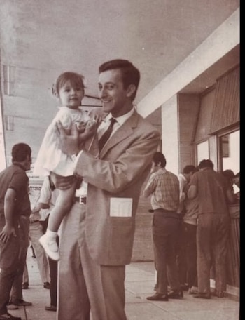 Un hombre vestido con traje sostiene a una niña pequeña con vestido blanco. Ambos sonríen. Varias personas se observan de fondo en un edificio