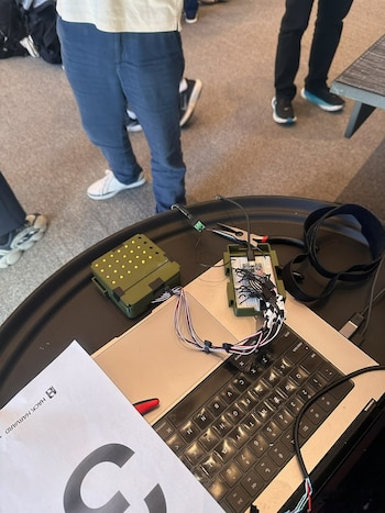 Computadora portátil con cables, pinzas, placas de circuitos y luces pequeñas sobre una mesa, junto a personas de pie
