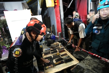Primer plano de rescatistas examinando más de diez teléfonos móviles quemados sobre una tabla de madera en un entorno de escombros y cinta de seguridad