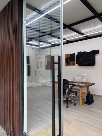 Puerta de vidrio con tirador negro y el texto Alterna, que muestra un interior con escritorio de madera, un portátil, sillas y decoraciones