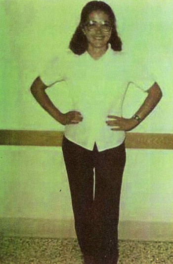Una mujer de cabello oscuro, blusa blanca de manga corta y pantalón oscuro posa con las manos en la cadera frente a una pared verde
