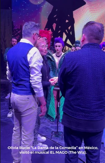 Olivia Bucio, de perfil con gafas y cabello rubio, conversa con un hombre de espaldas. Detrás, artistas con pelucas coloridas y disfraces en un escenario iluminado