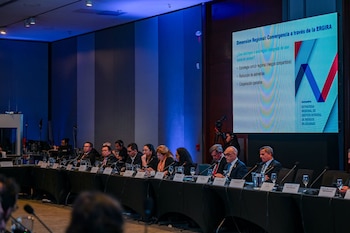 Varias personas sentadas en una mesa larga con micrófonos, vasos de agua y cartelitos. Detrás, una pantalla grande muestra una presentación