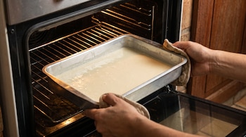 Dos manos con agarraderas sostienen y meten una bandeja metálica con masa líquida dentro de un horno abierto y encendido.