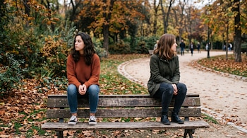Dos jóvenes están sentadas separadas en un banco de madera en un parque rodeadas de árboles y hojas secas de otoño.