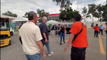 Shakira, con cabello rizado, top negro y jeans azules, camina por un estacionamiento. Hombres y un estadio con techo rojo son visibles al fondo