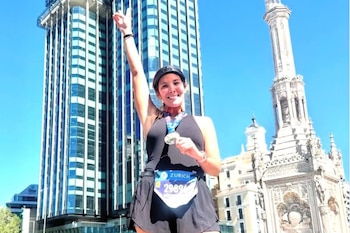 Una mujer con gorra negra, medalla al cuello y dorsal deportivo levanta el brazo frente a un rascacielos y un monumento