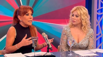Dos mujeres sentadas en una mesa con micrófono, vasos de agua y papeles. Una mujer pelirroja habla y una mujer rubia escucha. Fondo con diseño geométrico.
