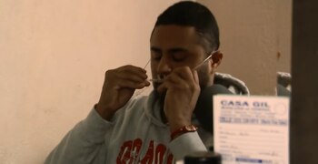 Un hombre con barba y bigote se inserta una cánula nasal. Viste una sudadera con capucha gris y un brazalete rojo. El fondo es una pared beige y un documento borroso
