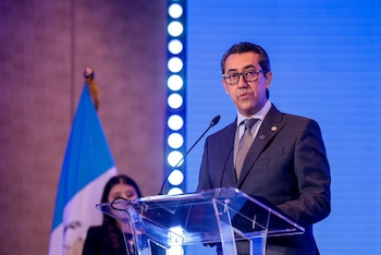 Hombre con traje y gafas en un podio transparente con micrófono; detrás, una mujer desenfocada y la bandera de Guatemala