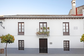 Fachada de una casa blanca de dos plantas con puerta oscura central, un balcón con plantas, cinco ventanas oscuras, tejado de tejas, dos naranjos y una farola