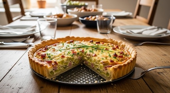 Tarta salada de puerro y jamón con una porción retirada, sobre un molde oscuro en una mesa de madera con cubiertos, platos y vasos.