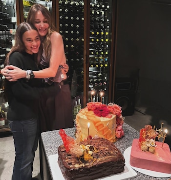 Verónica Lozano y una joven se abrazan sonriendo frente a una estantería de vinos. Sobre una mesa hay tres pasteles de cumpleaños con velas