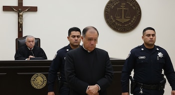 Juez con toga en estrado, hombre con sotana y rostro cubierto escoltado por dos policías, cruz y escudo de justicia al fondo en sala de tribunal.