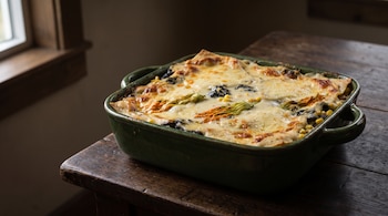 Lasaña mexicana en fuente de cerámica verde, con queso gratinado, huitlacoche, flor de calabaza y maíz, sobre una mesa de madera.