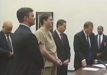 Un hombre con uniforme de prisión beige y esposas permanece de pie junto a varios hombres en traje y corbata en una sala judicial