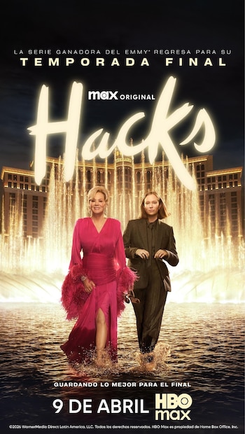 Jean Smart con vestido rosa vibrante y Hannah Einbinder con traje marrón caminan descalzas en el agua, con las fuentes del Bellagio y el hotel de fondo