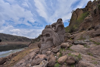 Escultura de roca gris con forma de calavera y figura alada en la orilla de un cuerpo de agua. Rocas, vegetación, montañas y cielo con nubes
