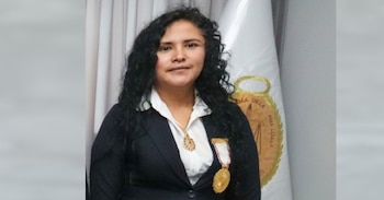 Retrato de la exfiscal Carmen López, una mujer con cabello oscuro y rizado, vistiendo un traje oscuro, camisa blanca y varias medallas. De fondo, una bandera