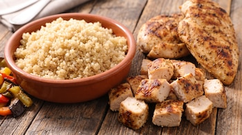 Un cuenco de barro con quinoa junto a trozos y filetes de pollo a la plancha en una mesa de madera con verduras.