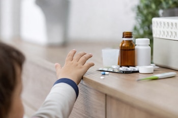 Mano de un niño alcanzando el borde de una mesa con frascos, tabletas de pastillas, un vaso medidor y un termómetro.