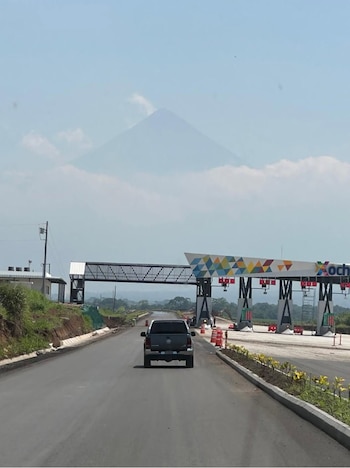 Camioneta oscura en carretera pavimentada hacia una plaza de peaje moderna con diseños geométricos. Un volcán grande y nebuloso se alza sobre las nubes en la distancia
