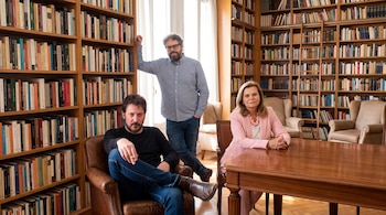 Tres personas en una biblioteca con libros en estanterías de madera, una mesa grande y sillones de cuero.