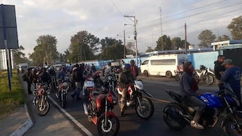 Varias personas a bordo de motocicletas rojas, azules y blancas se encuentran en una calle pavimentada cerca de una camioneta blanca