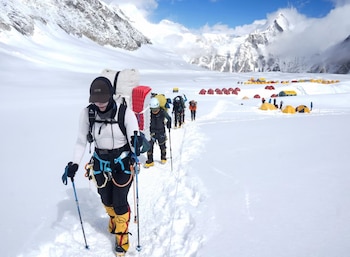 Un grupo de alpinistas con mochilas y bastones camina en fila por un sendero nevado. Al fondo, un campamento con tiendas rojas y amarillas y montañas nevadas
