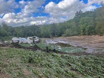 Un invernadero blanco colapsado yace en un campo cubierto de barro y vegetación aplastada. Hay un pequeño charco de agua y árboles verdes al fondo. El cielo está parcialmente nublado