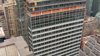 Una vista aérea de un edificio alto con múltiples ventanas y andamios naranjas en los pisos superiores, rodeado por otros edificios urbanos