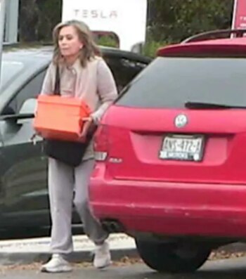 Mujer camina con caja naranja y bolso negro junto a auto rojo con matrícula mexicana. Se ve un cartel de Tesla al fondo