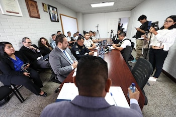 Personas vestidas de traje y policías uniformados sentados en una mesa de conferencia mientras dos personas graban con teléfonos