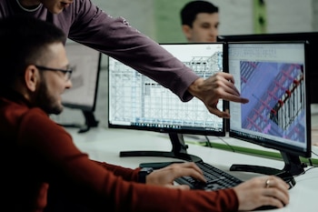 Dos personas observan monitores de computadora. Un hombre teclea, otro señala una pantalla. Los monitores muestran planos y modelos 3D de estructuras de edificios