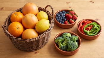 Una canasta de mimbre contiene naranjas, pomelos y limones. Tres cuencos de barro con arándanos, fresas, morrones rojos y verdes en rodajas, y floretes de brócoli.