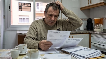 Hombre de mediana edad con jersey roto se rasca la cabeza y mira papeles en una mesa. Hay tres tazas sucias y un cartel "Se Alquila" en la ventana.
