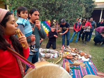 Grupo de personas en un espacio exterior. Una mujer y un hombre tocan zampoñas. Un hombre sostiene a un niño. Objetos, vasijas y alimentos en un paño en el suelo