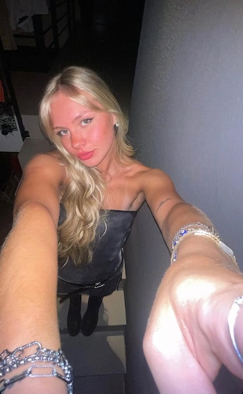 Mujer joven rubia con cabello largo, vestida de oscuro sin tirantes, aretes, collar y pulseras, incluyendo un tatuaje en brazo, en ambiente interior con escaleras