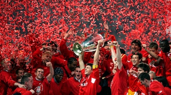 Jugadores de fútbol con camisetas rojas celebran en un estadio mientras el capitán levanta un trofeo de plata entre confeti rojo.