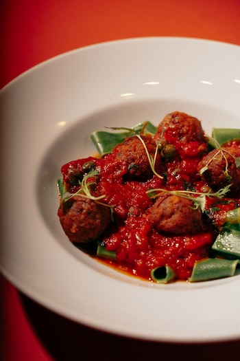 Primer plano de un plato blanco con pasta verde, albóndigas redondas, y una salsa de tomate rojo intenso, adornado con pequeñas ramitas verdes
