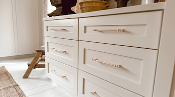 Mueble blanco con seis cajones y tiradores dorados, un taburete de madera, una alfombra tejida y un suelo de madera clara en una habitación iluminada.