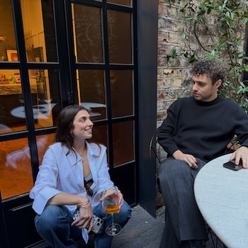 Un hombre y una mujer sentados en una terraza exterior. La mujer, en cuclillas, sonríe y sostiene una copa. El hombre, sentado en una silla, la mira