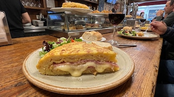 Primer plano de una porción de tortilla de patatas con jamón y queso fundido, acompañada de ensalada y pan, servida en una mesa de madera en un bar.